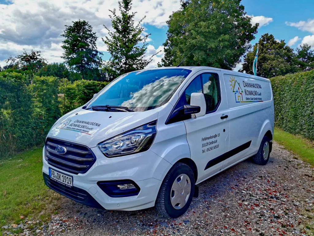 Ein weißer Transporter der Firma Danhausen & Kowalski steht auf einem Kiesweg umgeben von Bäumen und Sträuchern.