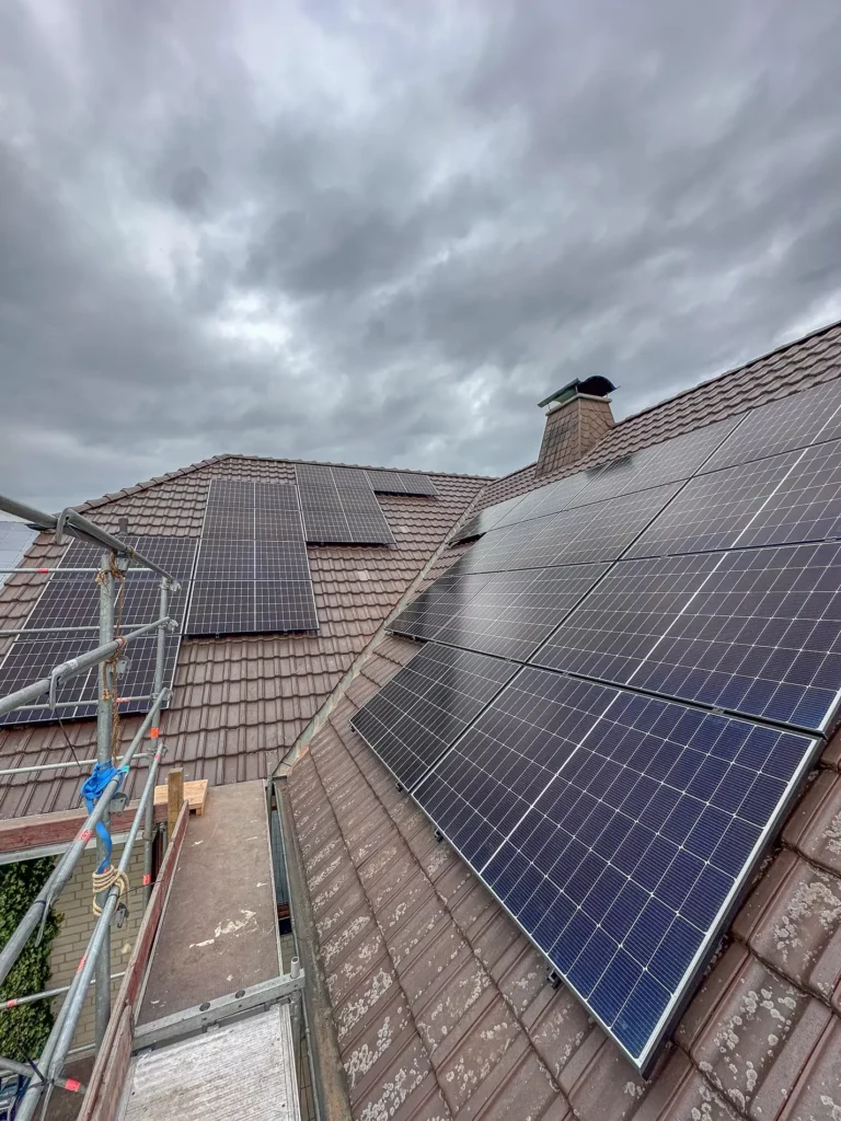 Auf einem Dach sind mehrere Solarpanels installiert, während ein Gerüst im Vordergrund zu sehen ist und der Himmel bewölkt ist.