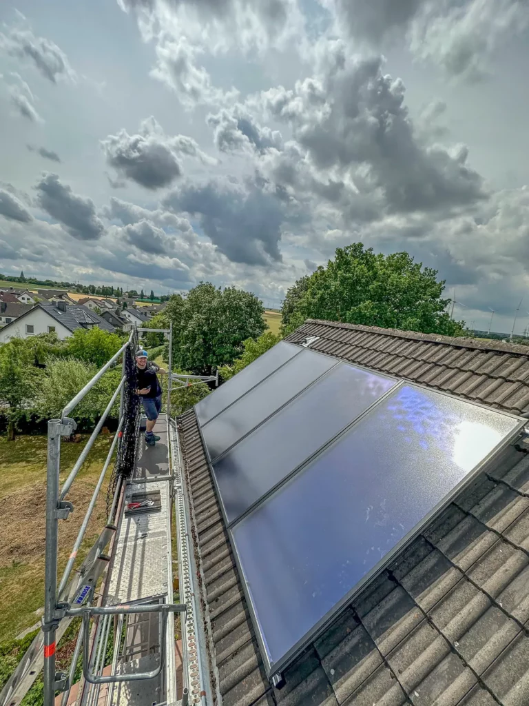 Ein Handwerker installiert Solarpanels auf einem Dach, während eine Wolkenlandschaft den Himmel über einer Wohngegend prägt.