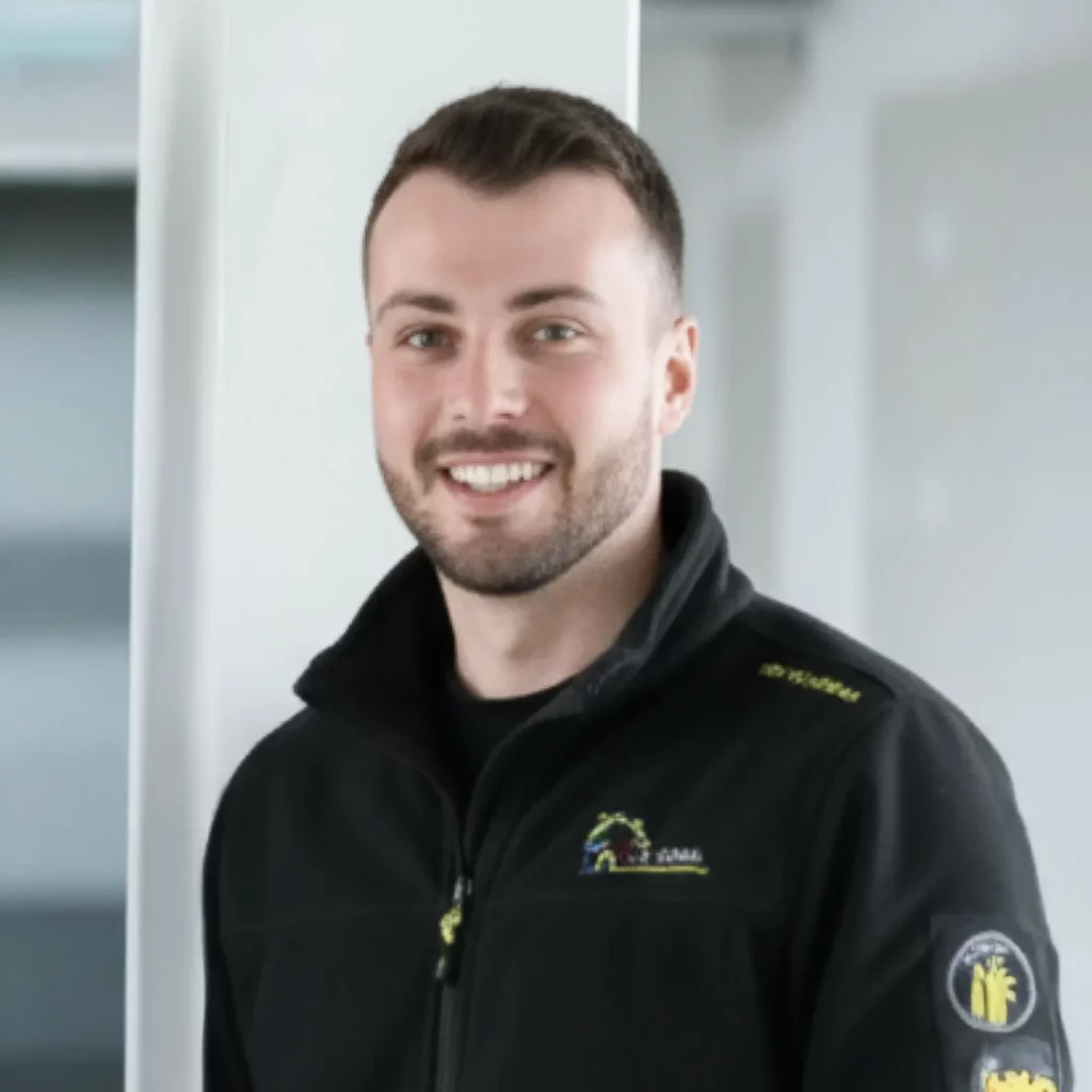 Junger Mann mit Bart lächelt in schwarzer Jacke vor modernem Hintergrund.