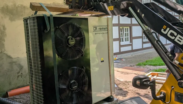 Ein Bagger hebt eine große Klimaanlage an, während sich ein historisches Fachwerkhaus im Hintergrund befindet.
