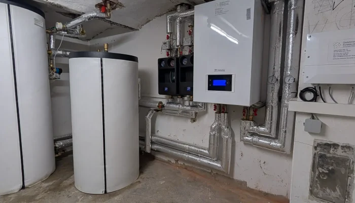 Der Raum zeigt zwei weiße Heizungsboiler und eine moderne Heizungssteuerung an der Wand, umgeben von isolierten Rohren.
