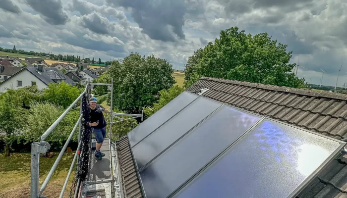Ein Handwerker installiert Solarpanels auf einem Dach, während eine Wolkenlandschaft den Himmel über einer Wohngegend prägt.